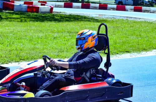 A go-kart driver in a colorful helmet races on a track, surrounded by green grass and red-and-white barriers.