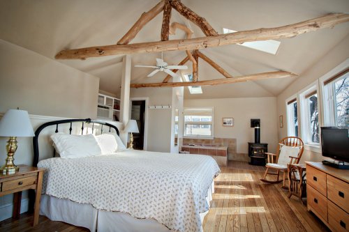 Bright bedroom with a wooden ceiling, large windows, a bed, and cozy seating, featuring natural light and warm decor.