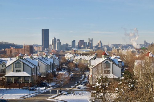 A snowy cityscape featuring houses in the foreground and a skyline with tall buildings in the background.