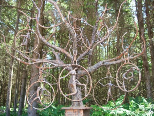 A metal tree sculpture with circular frames and figures hanging from its branches, set in a forested area.