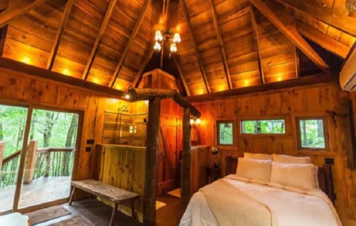Cozy wooden cabin interior with a bed, rustic decor, and large windows overlooking a forest. Warm lighting enhances ambiance.