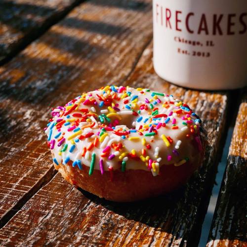 A colorful donut with white icing and sprinkles sits on a wooden table next to a coffee cup.