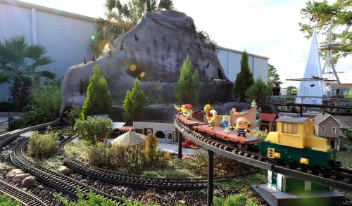 A miniature train travels along a track through a landscaped scene with trees, a mountain, and small buildings.