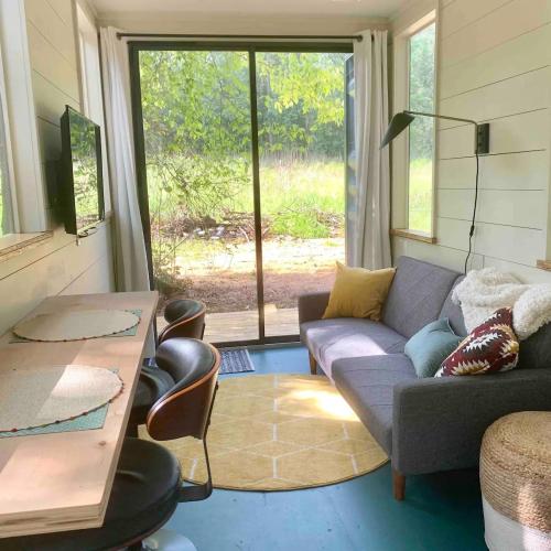 Cozy interior with a gray sofa, wooden dining table, and large windows overlooking a green outdoor space.