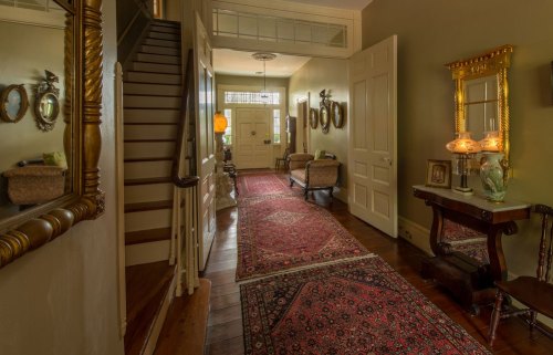 A spacious hallway with a staircase, vintage decor, mirrors, and a patterned rug, illuminated by natural light.