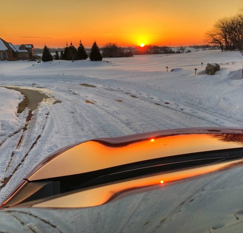 A snowy landscape at sunset, with the sun casting a warm glow on a car's hood and the road ahead.