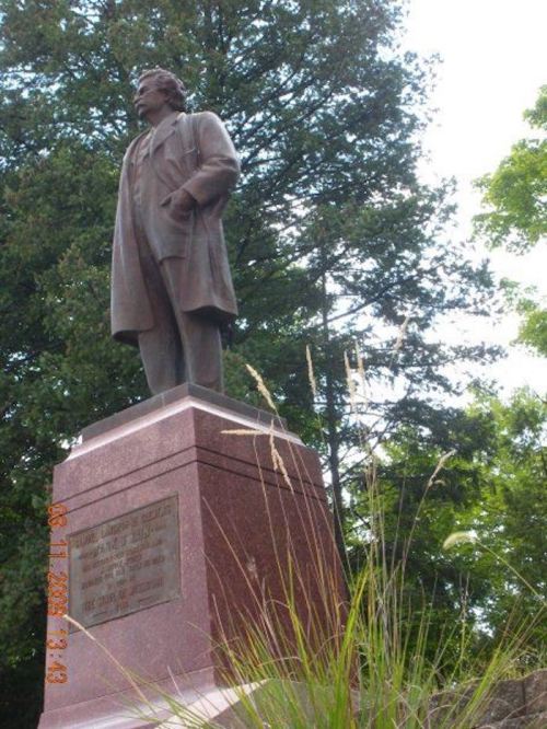 Statue of a man in a coat, standing on a pedestal surrounded by trees and grass.