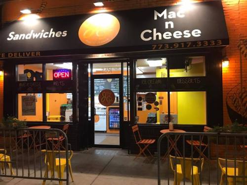 A brightly lit restaurant exterior featuring "Mac n Cheese" signage, outdoor seating, and an open sign at night.