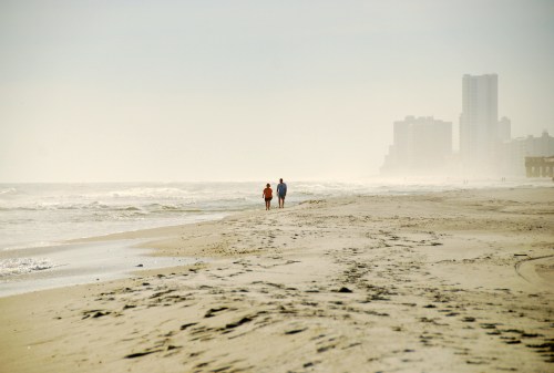 Two people walk along a sandy beach with gentle waves and a hazy skyline in the background.
