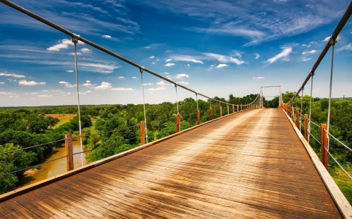 A wooden suspension bridge stretches over lush greenery and a river under a bright blue sky with scattered clouds.