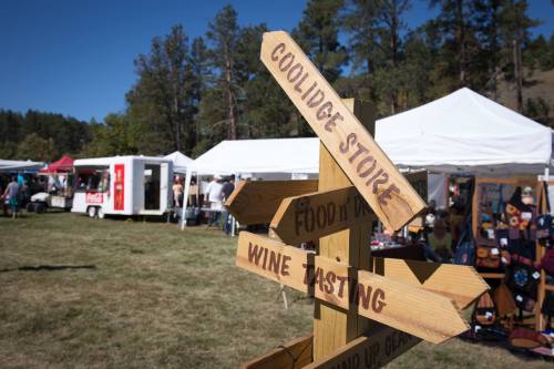 A wooden signpost with directions to various stalls at an outdoor market, surrounded by tents and trees.