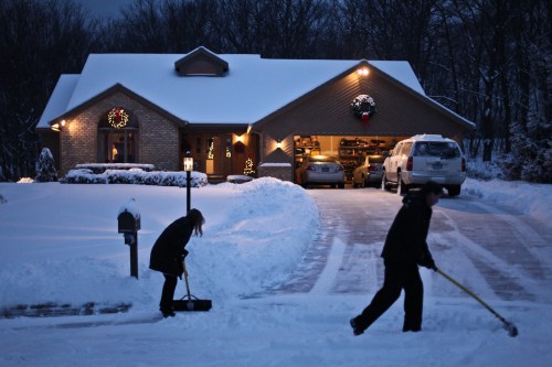 Two people shoveling snow in front of a house decorated for the holidays, with a snowy driveway and evening light.