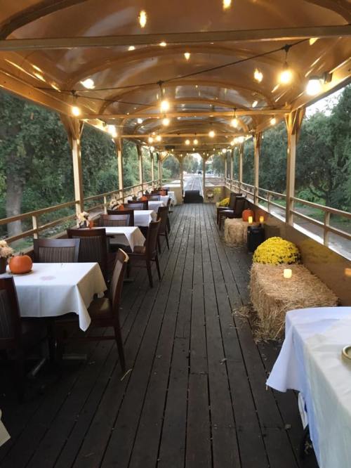 A cozy outdoor dining area with tables, string lights, and autumn decorations, surrounded by greenery.