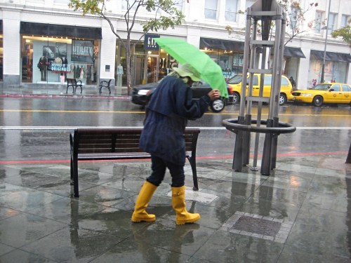 A person in yellow rain boots and a green umbrella walks along a rainy street, with taxis and shops in the background.