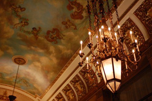 A decorative chandelier hangs from an ornate ceiling with painted cherubs and intricate gold detailing.