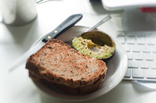 A plate with two slices of whole grain bread and half an avocado, next to a knife and a computer keyboard.