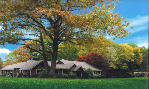 A large, brown house surrounded by colorful autumn trees and a green lawn under a clear blue sky.