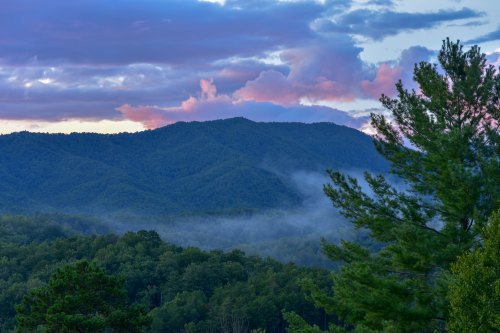 A serene landscape featuring rolling mountains, lush trees, and colorful clouds at sunset, with mist in the valley.