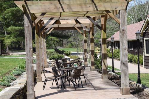 A wooden pergola with tables and chairs on a patio, surrounded by greenery and a pathway in a serene outdoor setting.