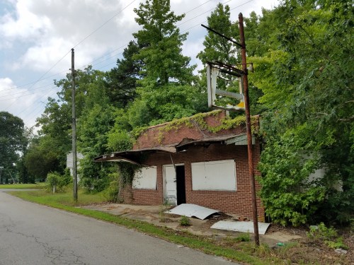 Abandoned brick building overgrown with vegetation, located on a quiet street, with boarded windows and a faded sign.