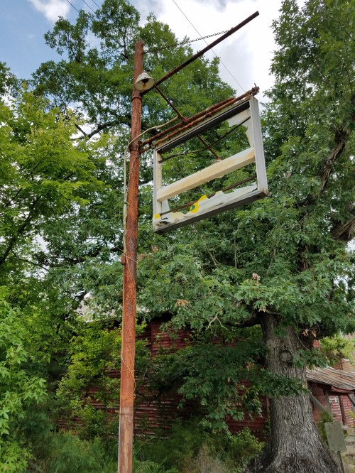 A rusty pole with a weathered, empty sign hangs amidst lush green trees.
