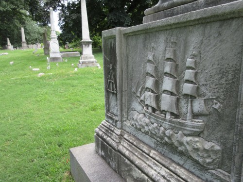 A close-up of a gravestone featuring a carved ship, with other gravestones visible in the background on a grassy area.