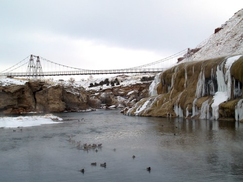 A snowy landscape featuring a frozen waterfall, a river with ducks, and a suspension bridge in the background.