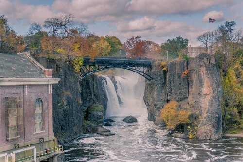A scenic view of a waterfall with a bridge above, surrounded by autumn foliage and a cloudy sky.