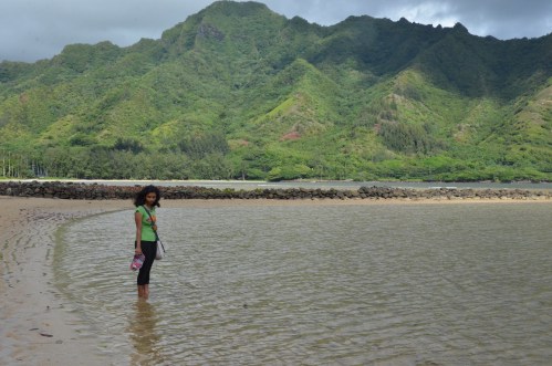 A girl stands in shallow water at a beach, with lush green mountains in the background under a cloudy sky.