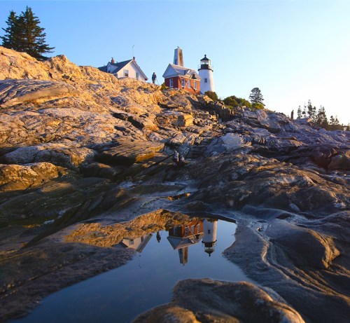 A lighthouse on rocky terrain reflects in a puddle at sunset, surrounded by trees and a clear blue sky.