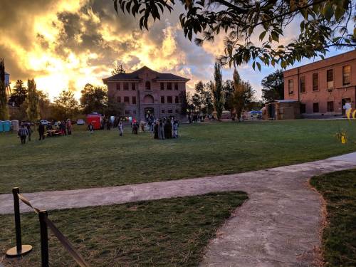 A grassy area with people gathered near buildings, under a dramatic sunset sky with clouds.