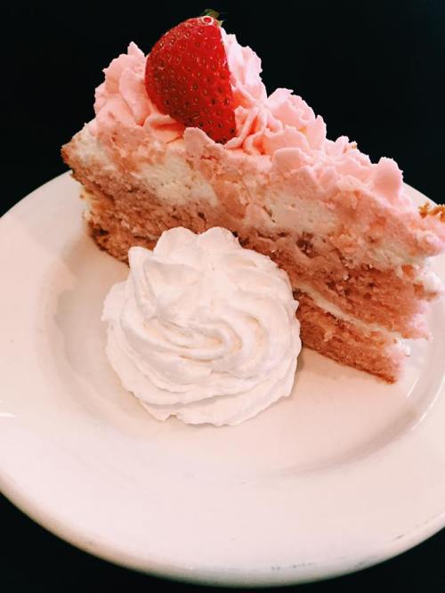 A slice of pink frosted cake topped with a strawberry, served with a dollop of whipped cream on a white plate.