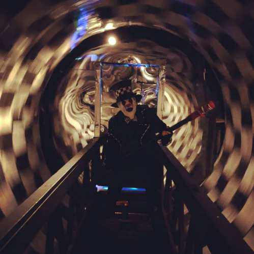 A person in a black outfit and hat stands on a bridge in a swirling, checkered tunnel, holding a large prop.