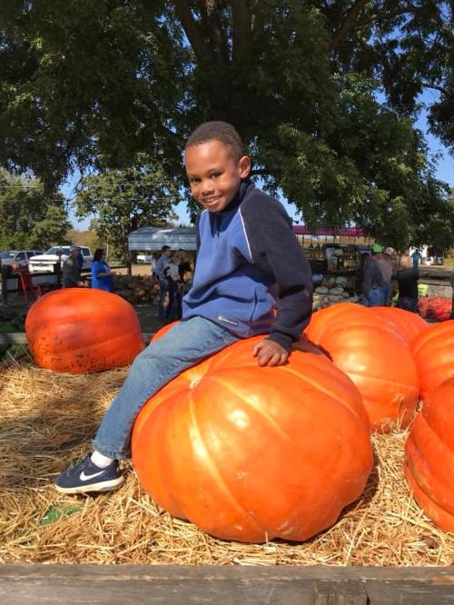 A smiling child sits on a large orange pumpkin at a pumpkin patch, surrounded by other pumpkins and greenery.