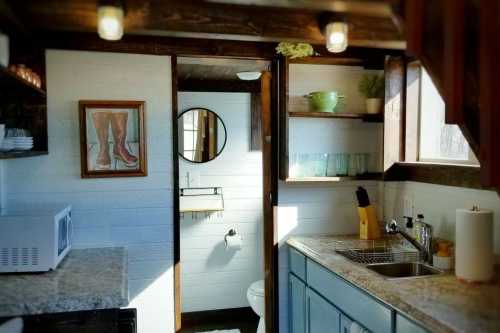 Cozy kitchen with a sink, microwave, and shelves, leading to a bathroom with a round mirror and rustic decor.