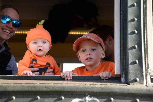 A baby in a pumpkin hat and a young boy in an orange cap peek out from a train window, smiling at the camera.