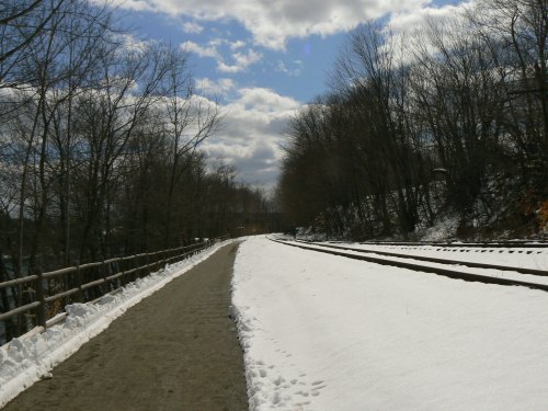 A snowy path alongside train tracks, with bare trees and a cloudy blue sky in the background.