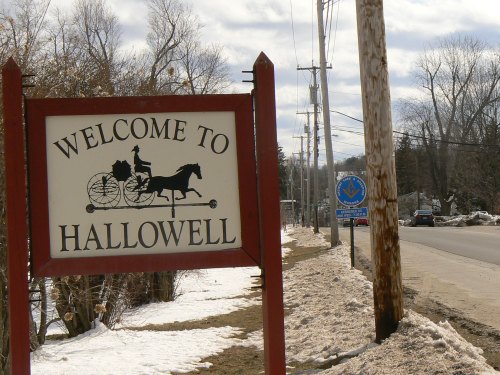Sign welcoming visitors to Hallowell, with a horse-drawn carriage graphic, alongside a snowy road and utility poles.
