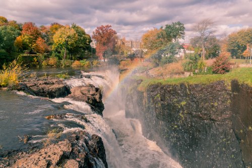 A scenic waterfall with a rainbow, surrounded by autumn foliage and a cloudy sky.
