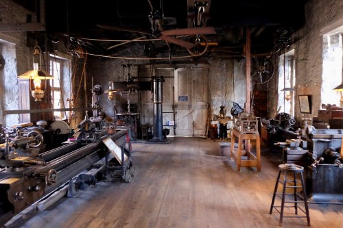 A vintage workshop filled with various machinery and tools, featuring wooden floors and large windows.