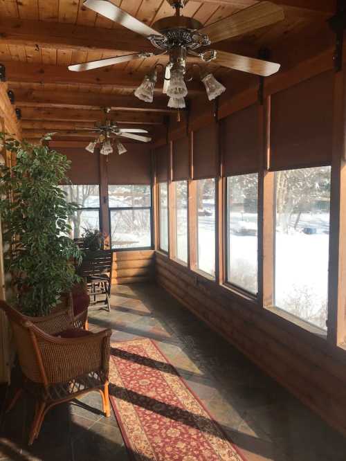 A cozy sunroom with wooden walls, ceiling fans, and large windows overlooking a snowy landscape.
