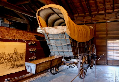 A vintage covered wagon displayed in a rustic wooden barn, with hay and historical images in the background.