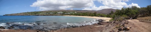 Panoramic view of a beach with clear water, rocky shore, and lush greenery under a partly cloudy sky.