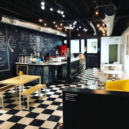 A modern café interior with black and white checkered floors, wooden tables, and a chalkboard wall. Brightly lit and inviting.