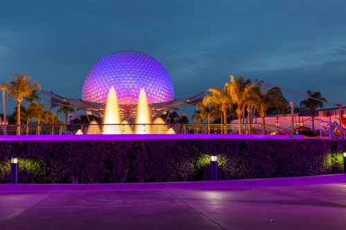 A futuristic dome structure illuminated in purple, surrounded by palm trees and a water feature at dusk.