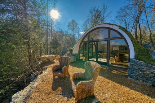 A modern, curved cabin surrounded by trees, featuring two wicker chairs on a gravel patio under a bright sun.