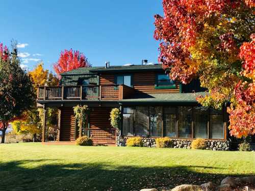 A log cabin surrounded by vibrant autumn foliage and a green lawn under a clear blue sky.