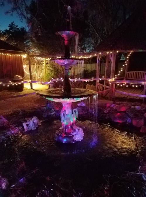 A colorful illuminated fountain at night, surrounded by twinkling lights and a serene garden setting.