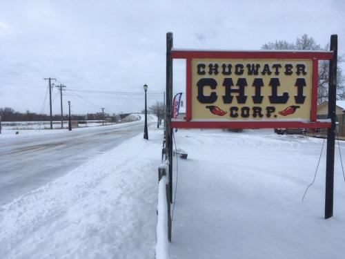Sign for Chugwater Chili Corp on a snowy road, with power lines and a cloudy sky in the background.
