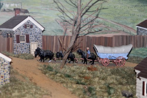 A miniature scene depicting a man guiding a horse-drawn wagon past stone houses and a barren tree.